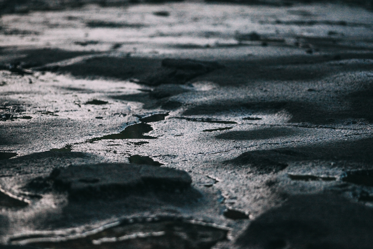 Close-up of a dark, cracked surface with shallow puddles, dimly lit and shadowed—evoking a mood of heaviness, isolation, and the absence of light or clarity.