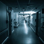 Dimly lit hospital hallway with closed doors on both sides, an empty wheelchair in the distance, and an illuminated EXIT sign above. The scene is quiet, sterile, and slightly eerie, evoking a sense of isolation or waiting.